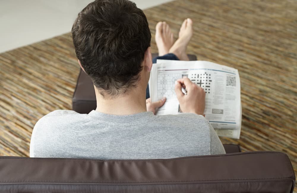 young man sitting on sofa and doing crossword puzzle in newspaper