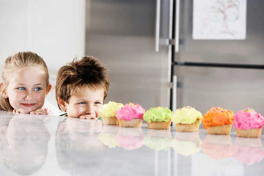 Young girl and boy peeking over counter at row of cupcakes